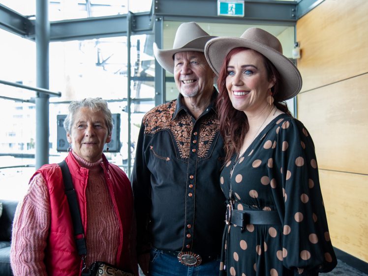 Image of Three people pose together indoors. An older woman in a red waistcoat stands next to an older man in a cowboy hat and embroidered shirt, with a younger woman in a polka-dot dress and hat smiling beside them.
