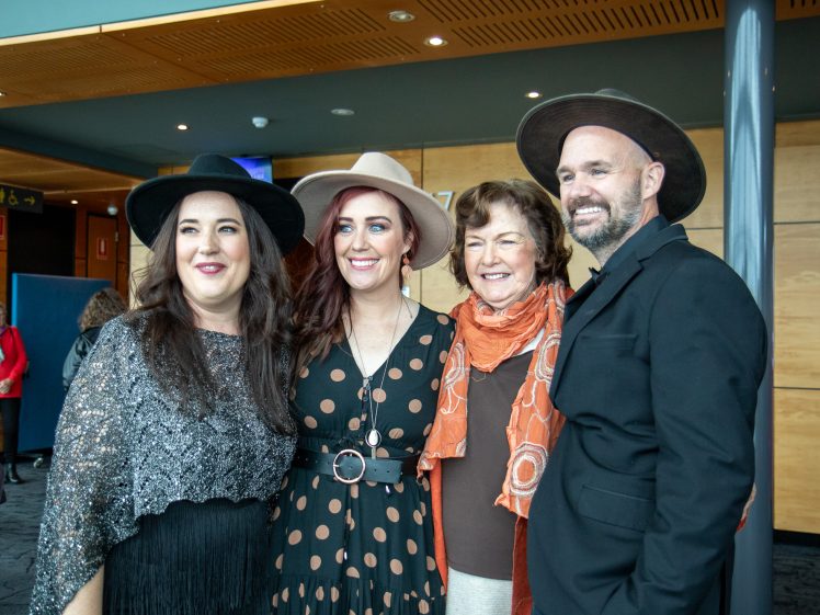 Image of Four people, three women and one man, stand together smiling. They are dressed in stylish outfits, with three of them wearing hats. They appear to be at an indoor event or gathering with others in the background.