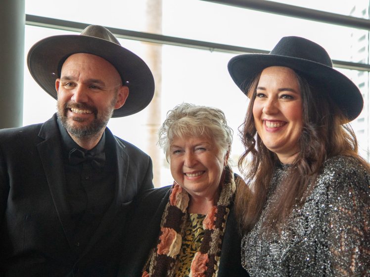 Image of Three people are standing together indoors, smiling. Two are wearing wide-brimmed hats and dark outfits, and the person in the middle is wearing a scarf with a floral pattern. Large windows are in the background.