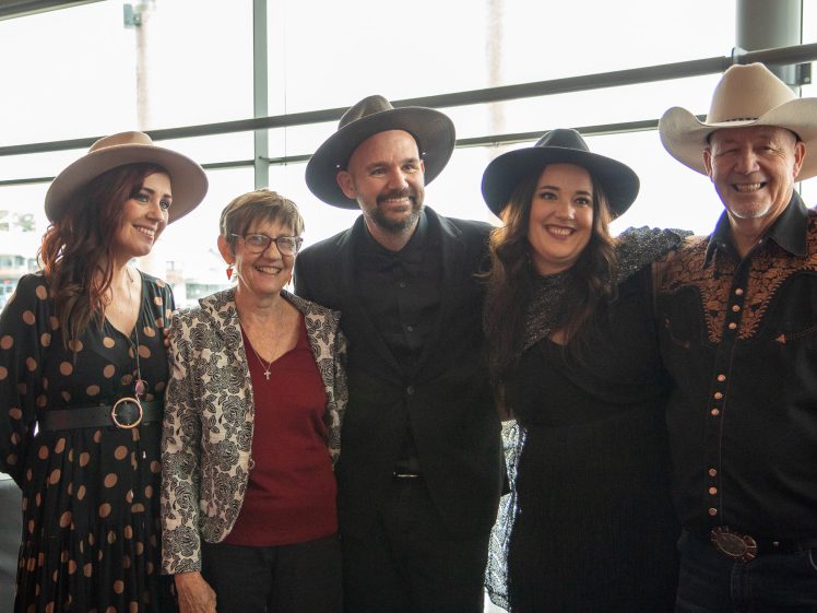 Image of Five people dressed in semi-formal and Western attire, including cowboy hats, stand together indoors by large windows, smiling and posing for a group photograph.