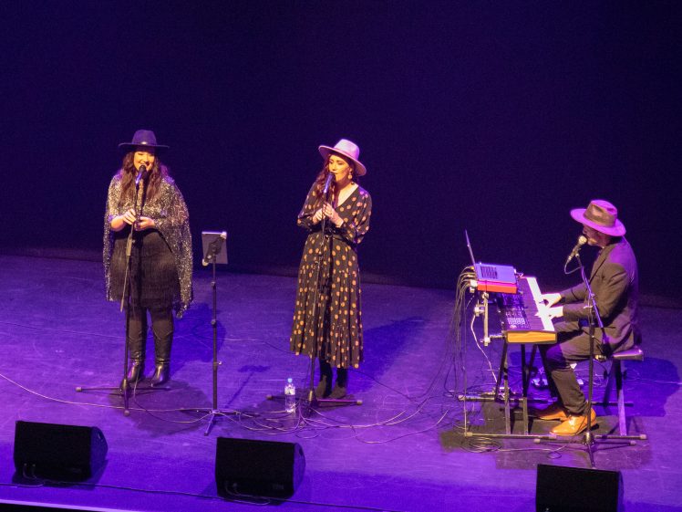 Image of Three musicians perform on a dimly lit stage—two women sing into microphones whilst a man plays a keyboard. All three wear hats and dark clothing, with music equipment and cables visible at their feet.