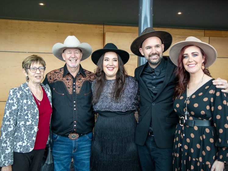 Image of Five adults stand together indoors, smiling at the camera. Two men wear cowboy hats and western-style shirts, while the three women wear hats and patterned outfits. The background features wood panelling and a glass column.