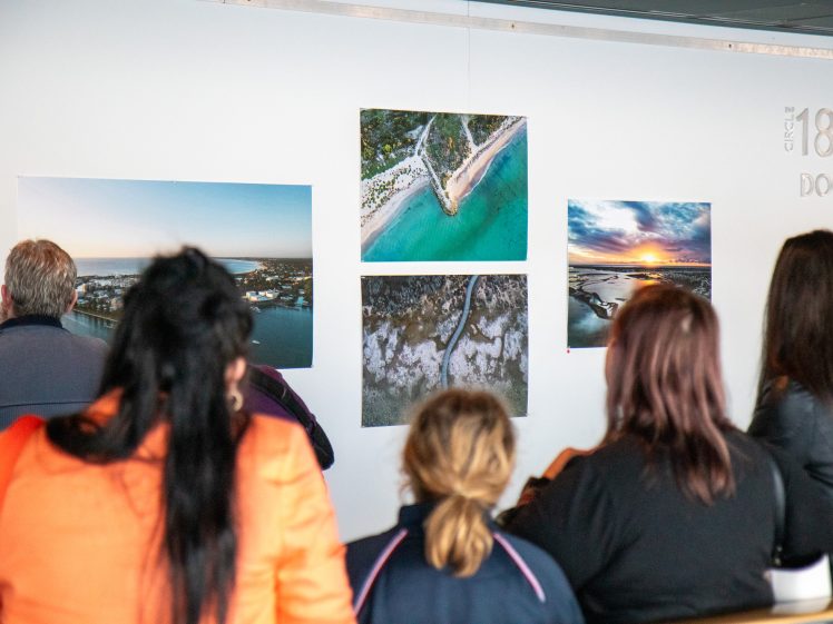 Image of A group of people stands in front of a wall displaying large aerial photographs of coastal landscapes at an indoor gallery or exhibition.