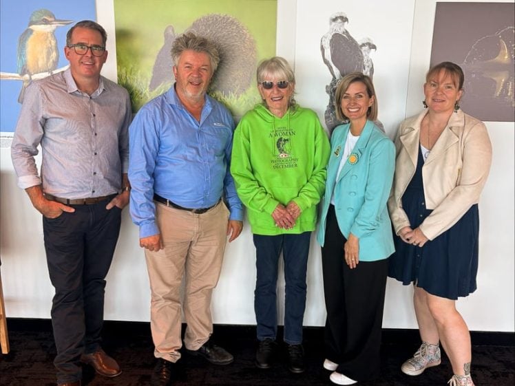 Image of Five adults stand indoors in front of a wall with wildlife photos, smiling at the camera. Three women and two men wear smart casual outfits, with one woman in a bright green hoodie.