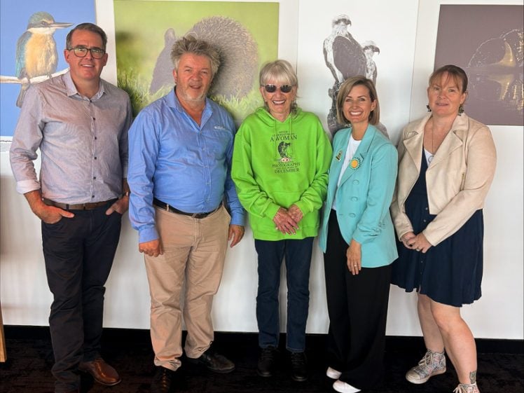 Image of Five adults stand in front of wildlife photographs on a white wall, smiling at the camera. They are dressed in business-casual and casual clothing, with one person wearing a bright green hoodie.