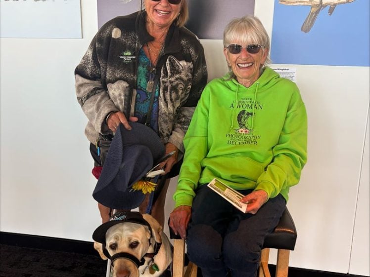 Image of Two smiling women wearing sunglasses pose in front of bird artwork; one stands holding a hat and a lead attached to a guide dog, while the other sits on a stool, holding a book. Both women have fair hair.