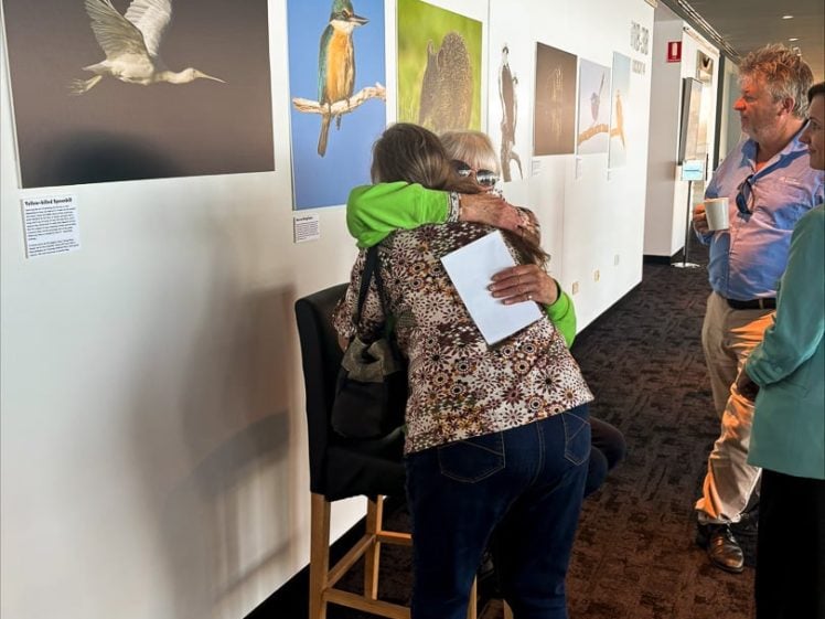 Image of Two women hug in front of a display of bird photographs on a gallery wall. One is sitting on a stool, whilst the other stands and embraces her. Two other people stand nearby, observing the scene.