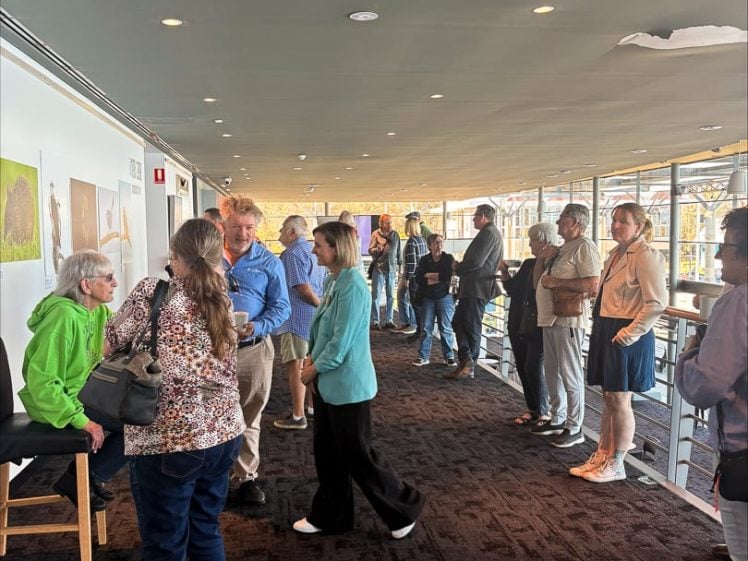 Image of A group of people stands in a corridor with large windows, some talking in pairs while others queue. Art hangs on the wall, and natural light fills the space.
