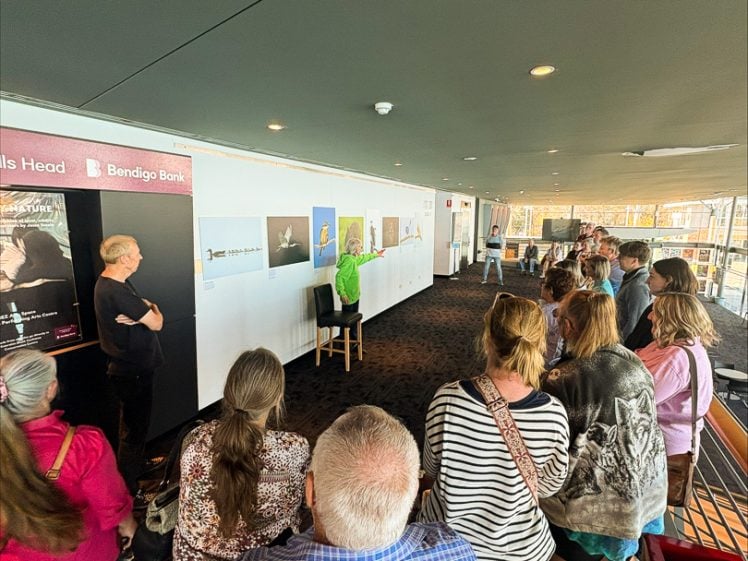 Image of A group of people stands in a carpeted room, facing a person seated on a stool near a wall with posters and a Bendigo Bank sign. The room has large windows letting in natural light.