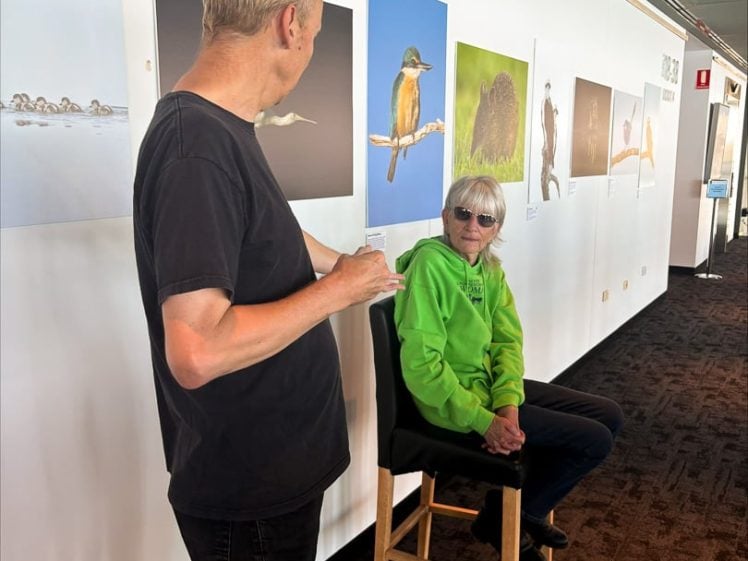 Image of A man in a black shirt talks to a woman in a bright green hoodie and sunglasses, who is seated on a stool in front of a white wall displaying bird photographs in a gallery setting.