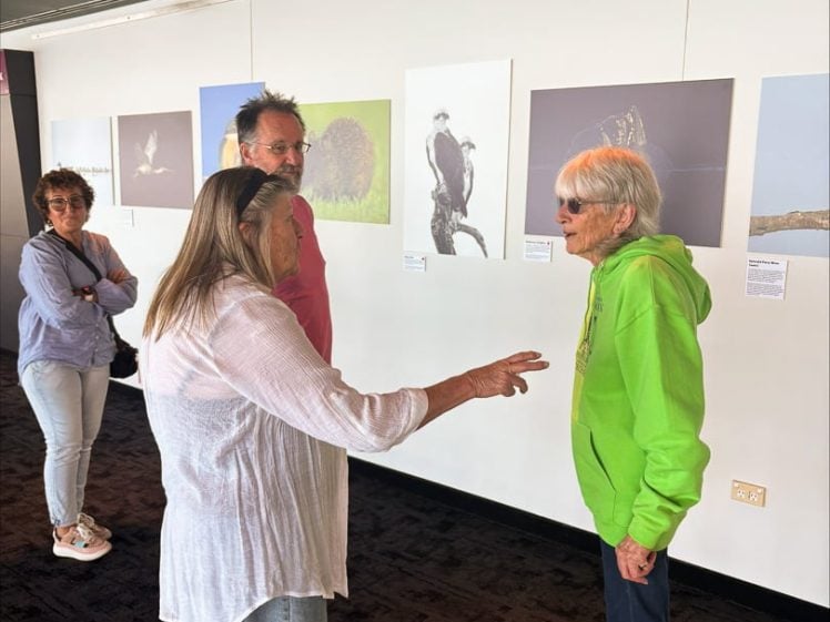 Image of Four adults stand in front of a wall displaying animal photographs, having a conversation. One woman gestures with her hand, whilst another in a green hoodie listens. Two others observe the scene.