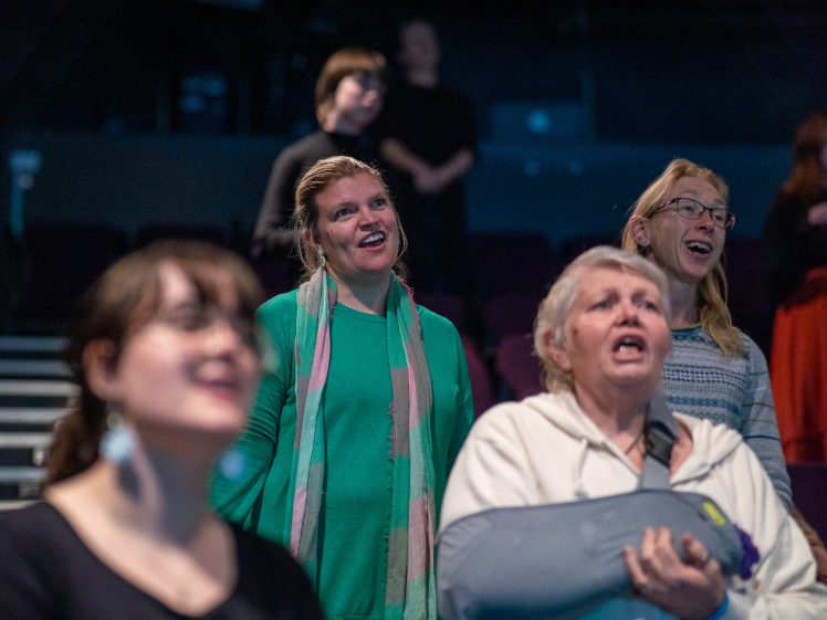 Image of A group of people, mostly women, stand closely together indoors, appearing to sing or speak passionately while facing forwards. Some are smiling, and one woman in front holds a grey cushion. The background is dimly lit.