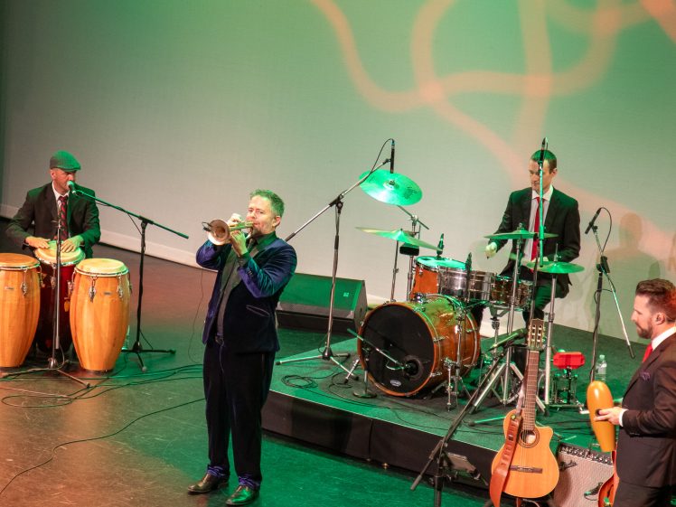 Image of A four-piece band performs on stage. One musician plays conga drums, another plays trumpet, a third is on drums, and the fourth holds a maraca. Various instruments and microphones are set up on stage under green lighting.