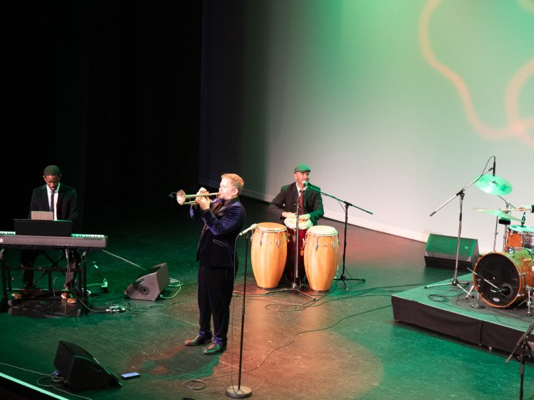 Image of A jazz band performs on stage. A keyboard player, trumpeter, percussionist with conga drums, and drummer are visible under green and white stage lighting. The trumpeter stands at the front, playing his instrument.