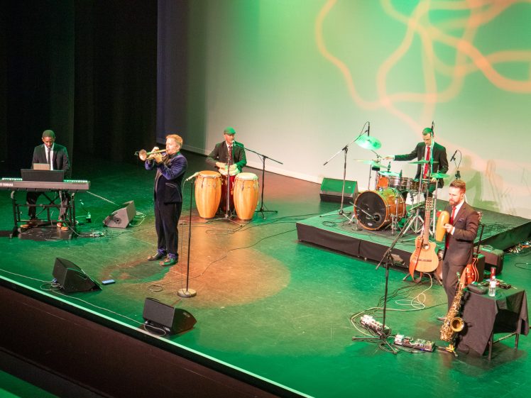 Image of A jazz band performs on stage with a keyboardist, trumpeter, conga player, drummer, and saxophonist. Green and yellow lighting patterns are projected on the backdrop.
