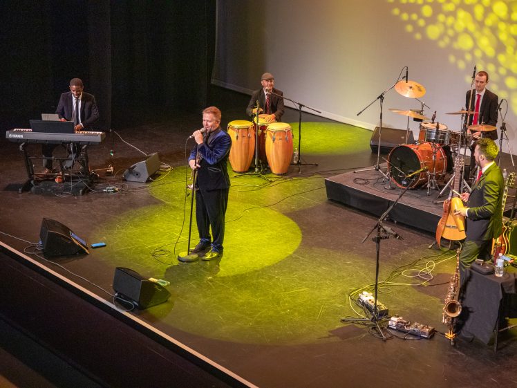 Image of A band performs on stage with a keyboard player, a singer, two percussionists playing drums and congas, and a guitarist; yellow stage lights create a lively atmosphere.