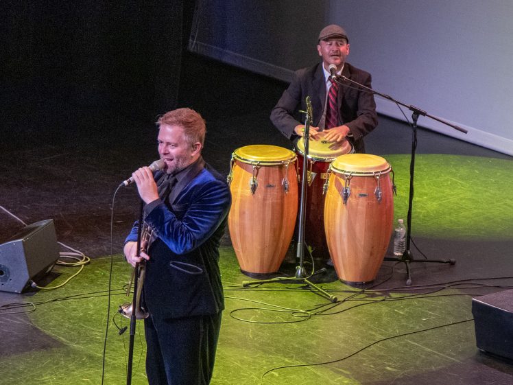 Image of A man in a blue suit sings into a microphone while holding a trumpet, and another man in a hat and suit plays conga drums onstage under colourful stage lights.