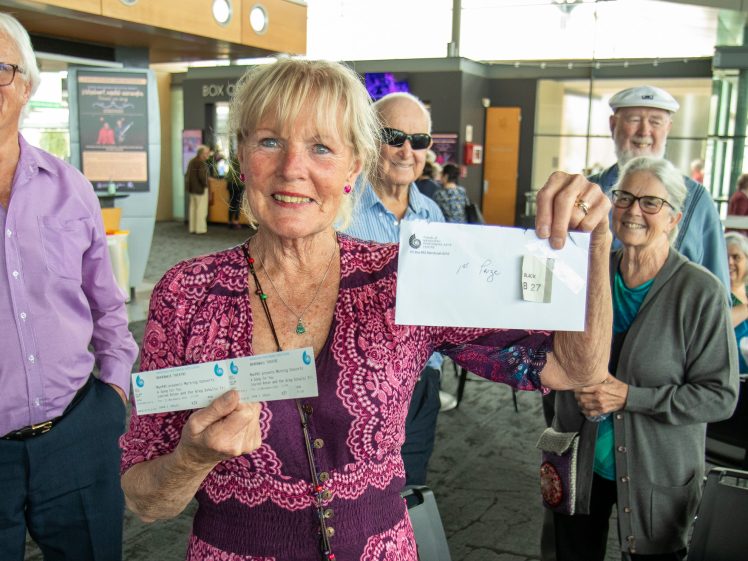 Image of An elderly woman smiles and holds up an envelope and tickets in a bright indoor setting, surrounded by other pensioners who are also smiling and looking at the camera.