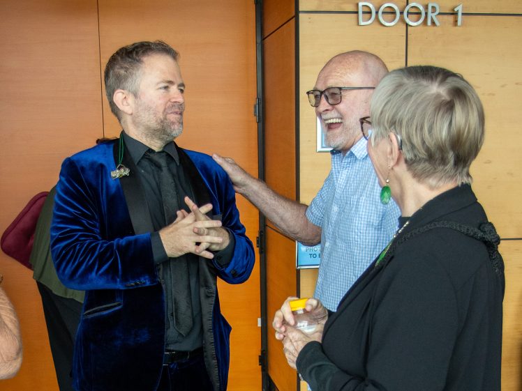 Image of A man in a blue velvet jacket talks and smiles with an older man and woman, all appearing happy and engaged, standing indoors near a wooden wall labelled DOOR 1.