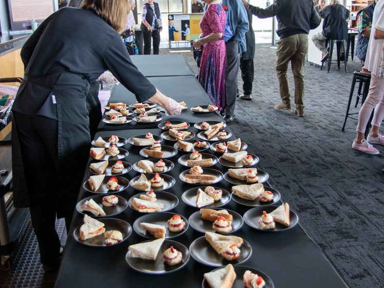Image of A person arranges small plates of appetisers on a long table, while people in semi-formal attire gather and converse in a bright, modern indoor space.