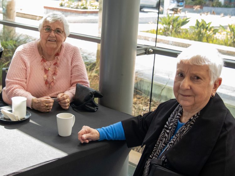 Image of Two elderly women sit at a table in a bright indoor space with large windows. Both have short white hair; one wears a pink jumper and spectacles, the other a black jacket. Coffee cups are on the table.
