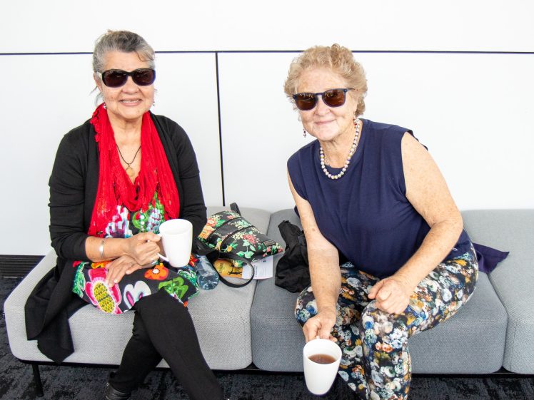 Image of Two older women sit on a grey sofa, smiling, each holding a mug. Both wear sunglasses and colourful outfits. A floral bag rests between them. The background is plain white with a thin black line.