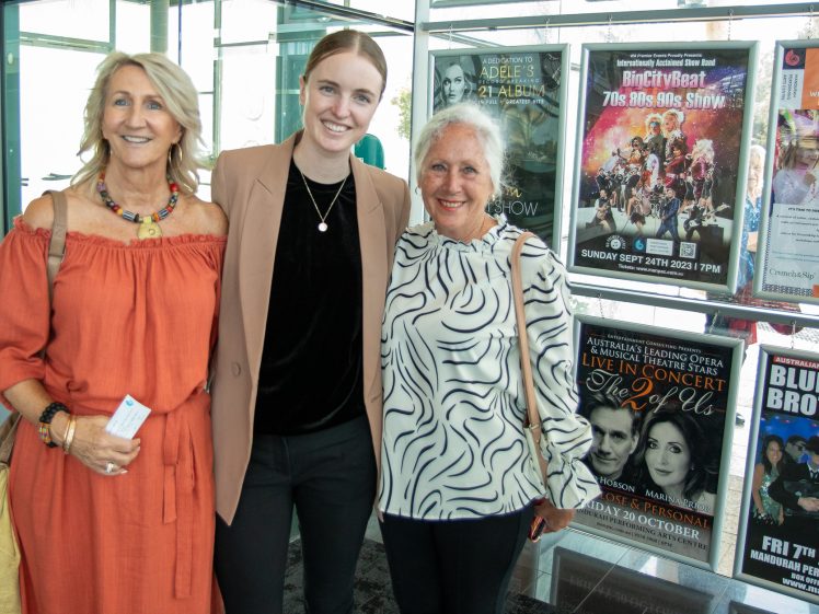 Image of Three women stand smiling together in front of a wall of colourful event posters, including ones for concerts and shows. Two women wear patterned tops, and one wears an orange dress. They appear to be at an event venue.