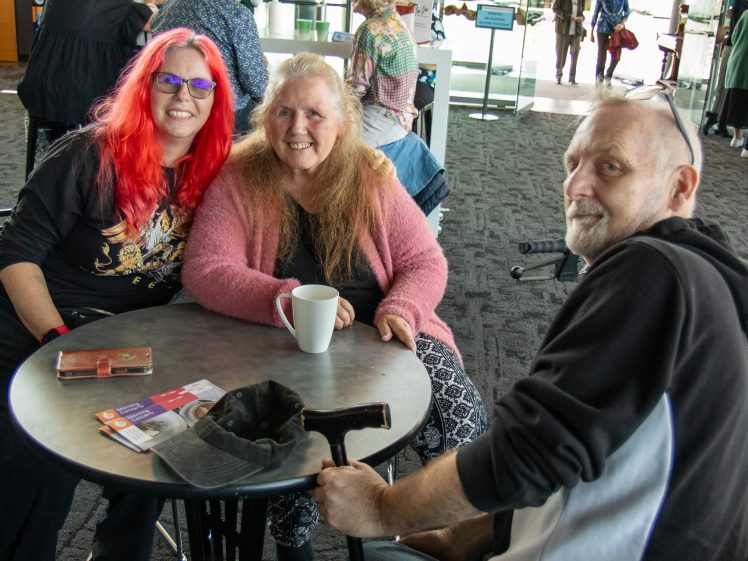 Image of Three smiling adults sit around a circular table in a public indoor space. Two women sit side by side; one has bright red hair and glasses, the other has long blonde hair and a pink jumper. A man with a walking stick sits opposite them.