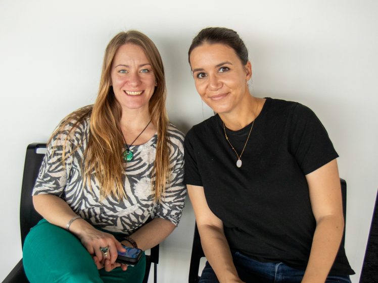 Image of Two women sitting side by side on chairs, smiling at the camera. One has long blonde hair, a patterned shirt, and green trousers; the other has dark hair tied back, a black t-shirt, and blue jeans. White background.