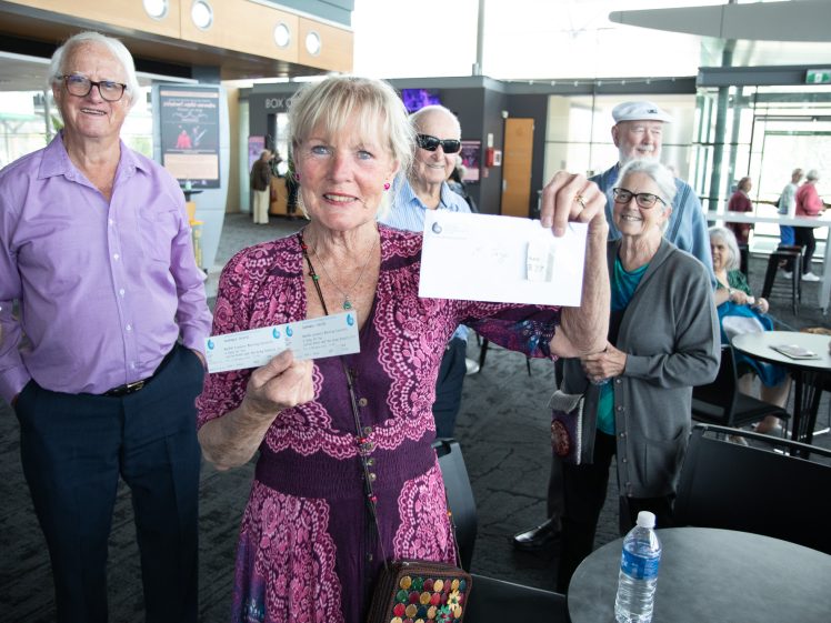 Image of An older woman in a pink dress smiles and holds up event tickets and a piece of paper, while a group of older adults stand and sit behind her in a bright, modern foyer.