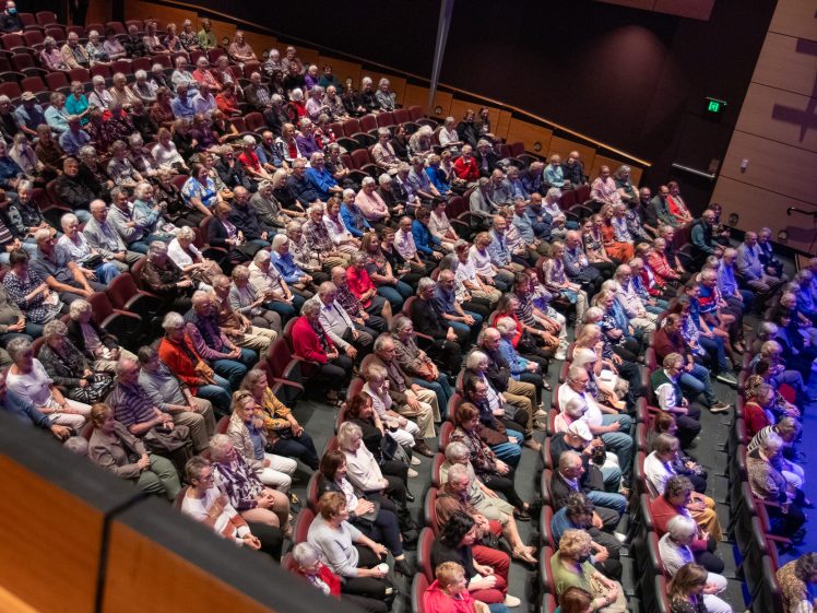 Image of A large audience sits closely together in a theatre, facing the stage. The seats are mostly filled, and people appear attentive, suggesting they are watching a performance or presentation.