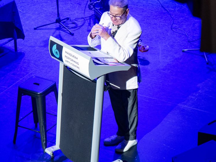 Image of An older person in a white jacket stands at a lectern on a blue-lit stage, holding a microphone and speaking. A black stool, musical equipment, and cables are visible in the background.
