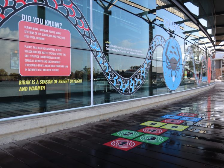 Image of A glass wall with Aboriginal artwork, informational signs about Birak season, and colourful hopscotch patterns on a wet wooden pathway. Reflections are visible on the glass and floor.