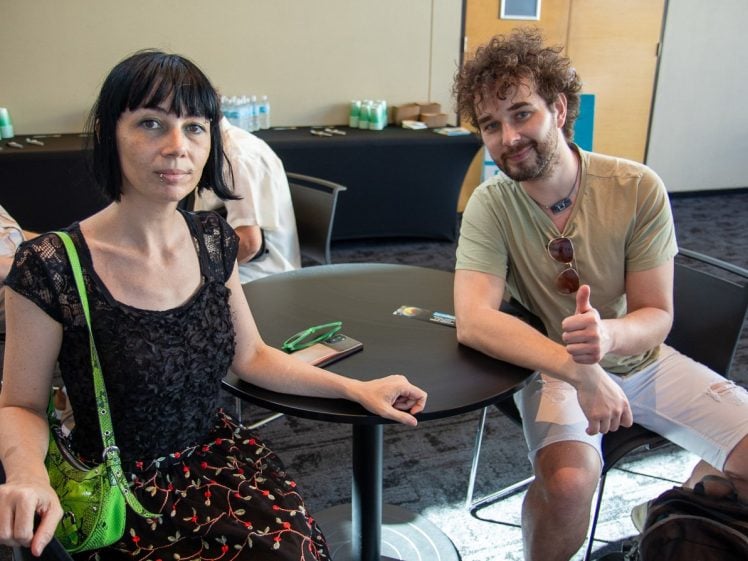 Image of A woman and a man sit at a round table indoors. The woman has dark hair and wears a black dress with a green bag, while the man, with curly hair and sunglasses, wears a beige shirt and shorts, giving a thumbs up and smiling.