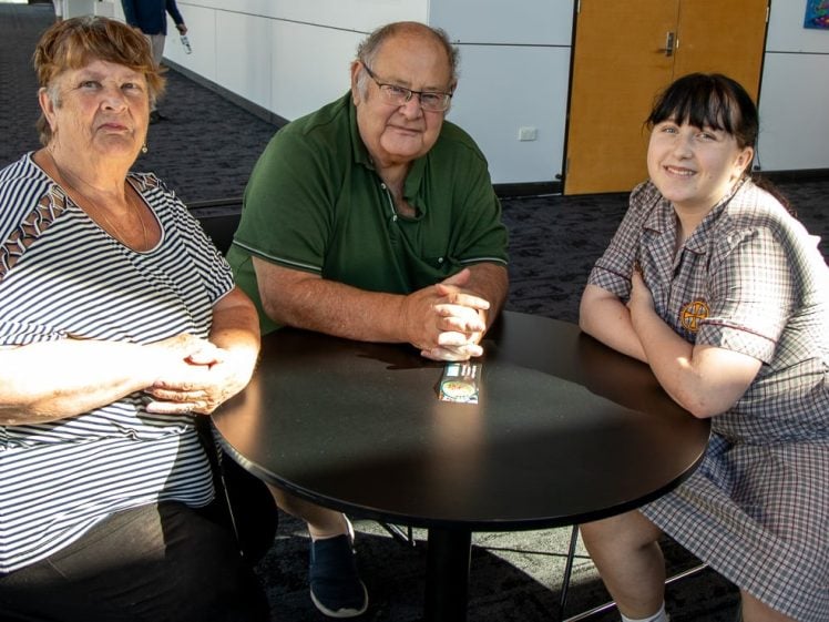 Image of Three people—an older woman, an older man, and a young woman in a chequered uniform—sit together at a round black table in a brightly lit indoor setting, smiling at the camera.