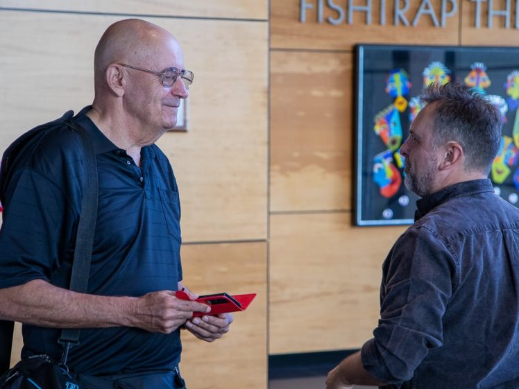 Image of Two men are talking in front of a wall with Fishtrap Theatre signage and colourful artwork. One man is holding papers and wearing glasses and a rucksack; the other is facing away from the camera.