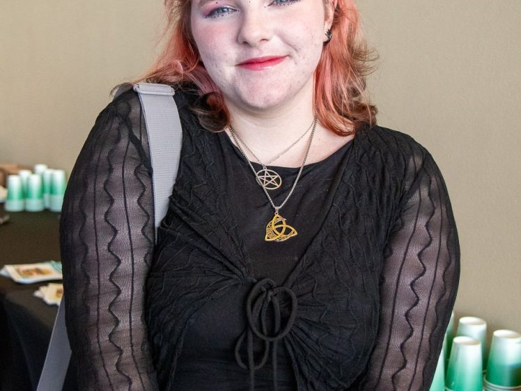 Image of A person with wavy reddish hair, wearing a black outfit and layered necklaces, stands smiling in front of a table with teal and white cups and snacks against a beige wall.