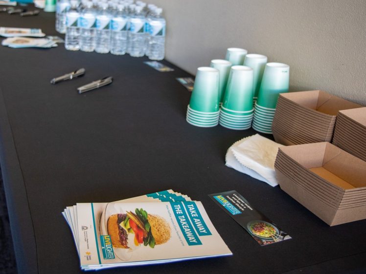 Image of A table displays bottled water, stacked green paper cups, cardboard food containers, serviettes, brochures with a burger image, and pens, set up for an event or meeting.
