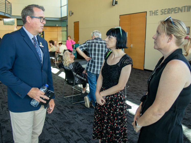 Image of A man in a suit holding a water bottle talks to two women in a lobby area, whilst several people sit or stand in the background near wooden doors labelled THE SERPENTINE.