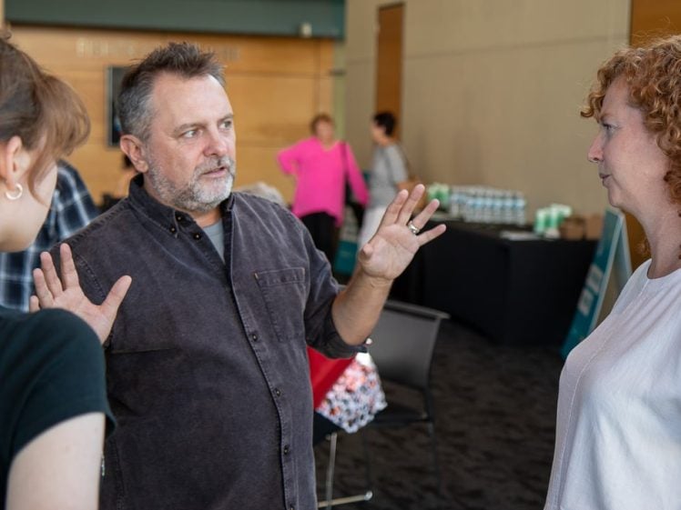 Image of A man gestures with his hands whilst talking to two women indoors. Other people and a table with drinks are visible in the background.