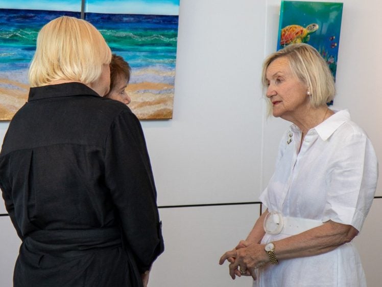 Image of Three women are standing and talking in an art gallery. Two women face away from the camera, while the third, wearing a white dress, faces them and speaks. Colourful ocean-themed paintings hang on the wall behind them.