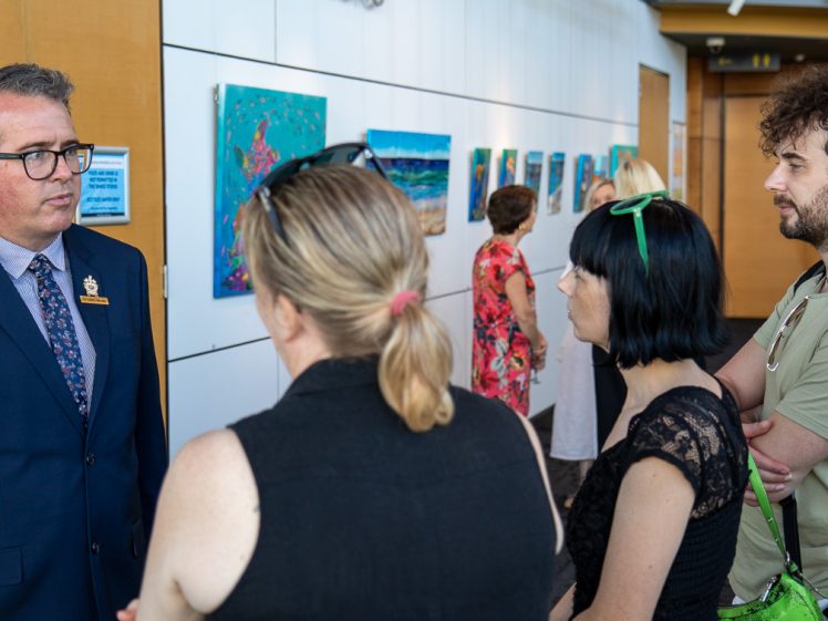 Image of A man in a suit speaks to three people in a gallery with colourful artwork on the walls. Other visitors view the art in the background.