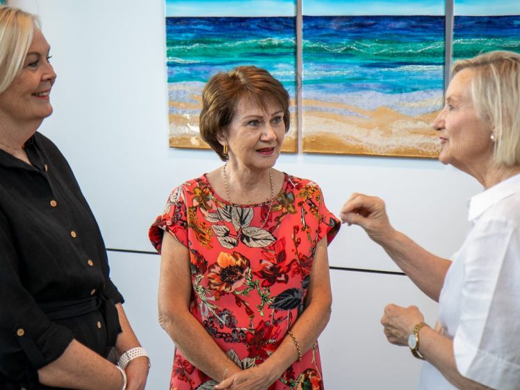 Image of Three women stand together indoors, engaged in conversation. Two wear dark and white dresses, while the third, in the centre, wears a red floral dress. A vibrant ocean-themed painting hangs on the wall behind them.