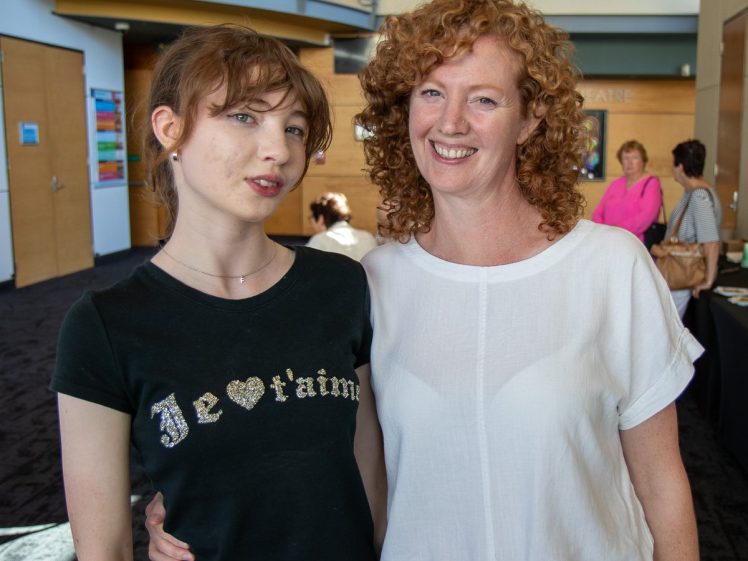 Image of Two women stand close together indoors, smiling at the camera. One has long curly red hair and wears a white blouse; the other has light brown hair and wears a black “Je t’aime” T-shirt. People and modern décor are in the background.