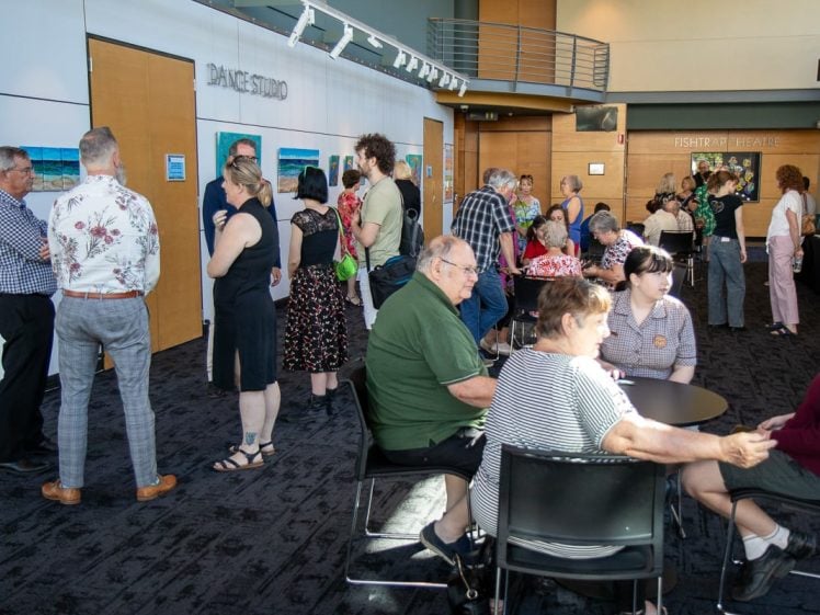 Image of A group of people socialise, chat, and sit at tables in a bright indoor event space with artwork on the walls and a sign reading DANCE STUDIO. Some people stand while others are seated, enjoying the gathering.