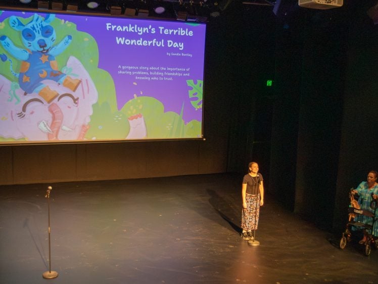 Image of Two people stand on a dimly lit stage. Behind them is a large screen displaying the title Franklyn’s Terrible Wonderful Day with colourful cartoon animals and a story description. A microphone stands on the left.