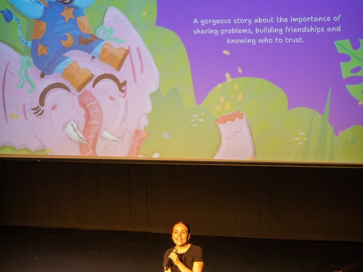 Image of A woman stands on stage with a microphone, smiling up at a large screen displaying a colourful book cover titled Franklyn’s Terrible Wonderful Day with an illustrated blue cat riding a pink elephant.