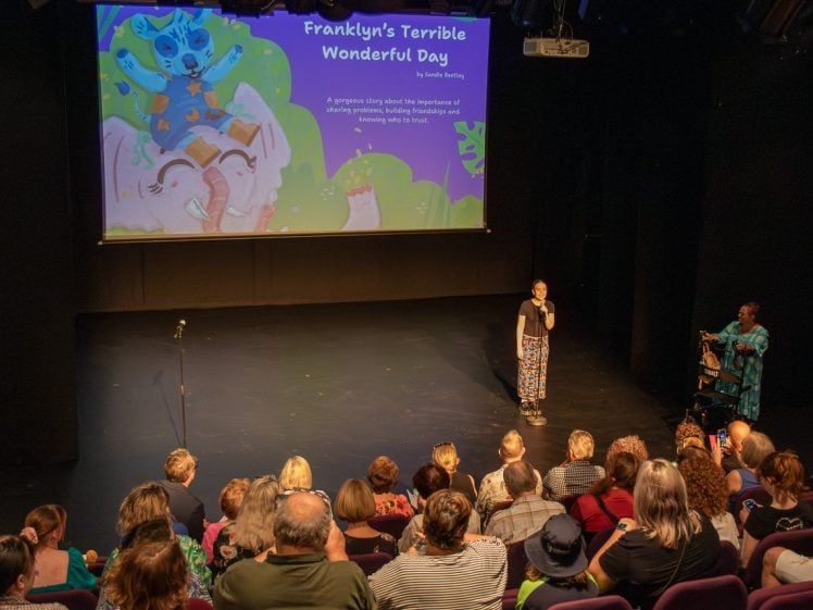 Image of A person stands on stage in front of an audience, presenting Franklyn’s Terrible Wonderful Day, a children’s story book. A bright, colourful illustration is projected behind them, while a sign language interpreter stands at the side.
