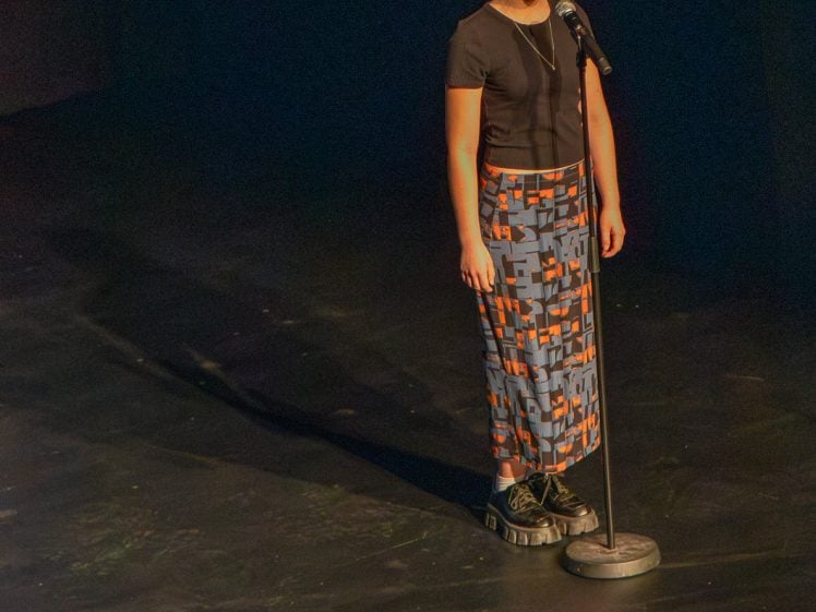 Image of A person stands alone on a dimly lit stage, singing or speaking into a microphone. They wear a black top, geometric patterned skirt, and black platform shoes. A shadow stretches behind them.