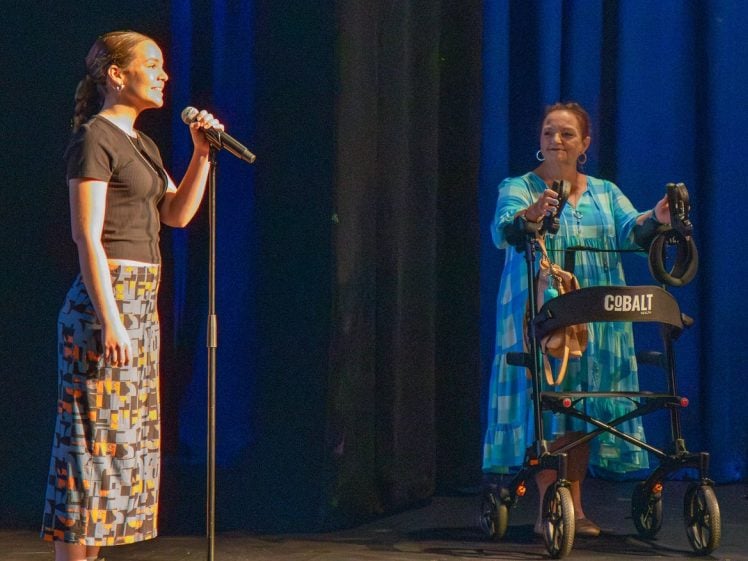Image of A woman stands on stage speaking into a microphone whilst another woman in a blue dress stands beside her holding a Cobalt walking frame, both illuminated by stage lights against a dark background.